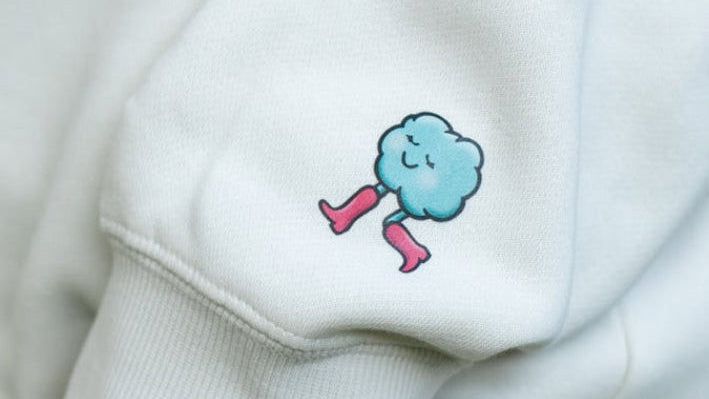 Cream sweatshirt with a blue cloud character wearing pink shoes on a cream background