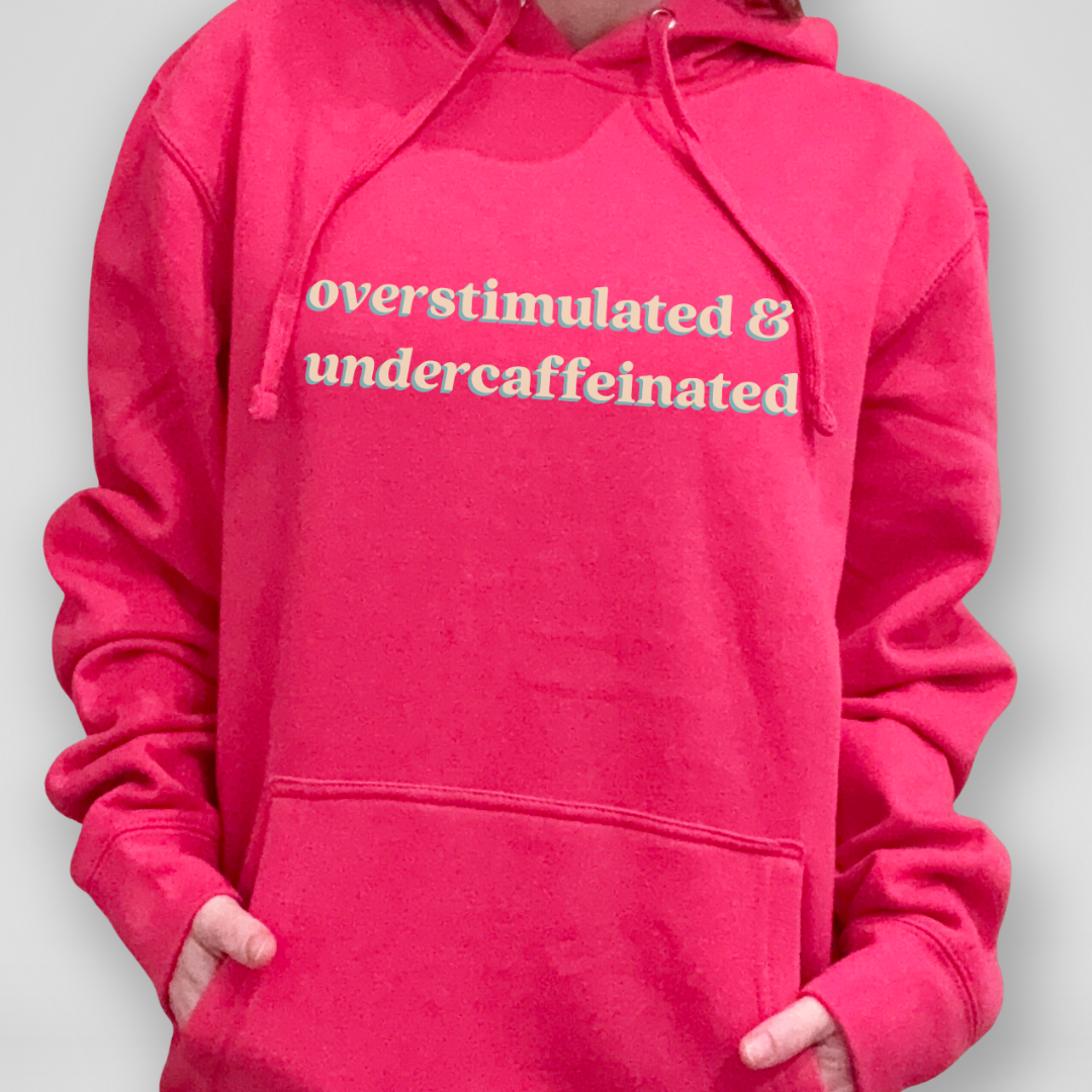 Person wearing a pink hoodie with text 'overstimulated & undercaffeinated' on a plain background