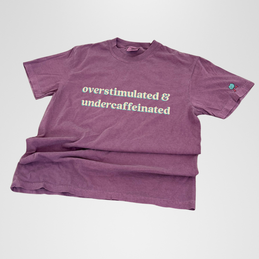 Purple t-shirt with text 'overstimulated & undercaffeinated' on a light gray background