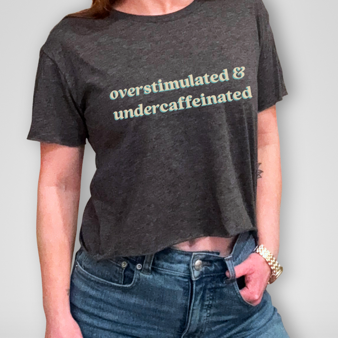 Person wearing a dark gray t-shirt with 'overstimulated & undercaffeinated' text, paired with blue jeans on a plain background.