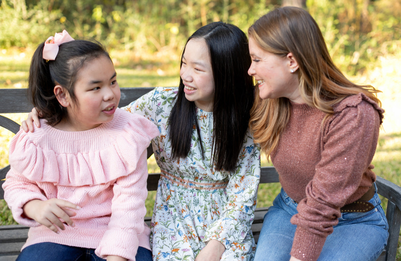 Three young women sitting on a bench outdoors, smiling and laughing.