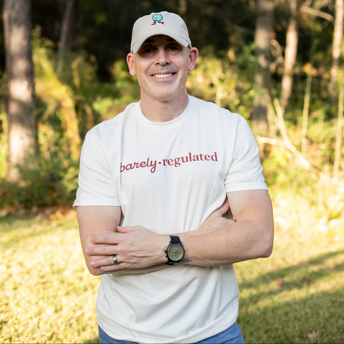 Man wearing a cream t-shirt with 'barely regulated' text and a cap, standing outdoors with trees in the background.