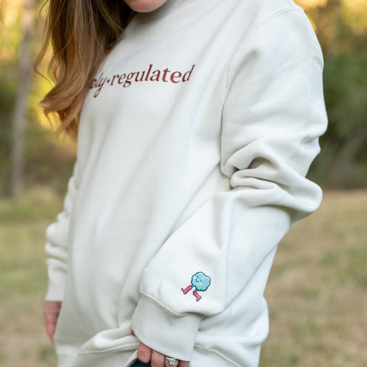 Person wearing a cream sweatshirt with 'Barely Regulated' text and a small cloud design outdoors