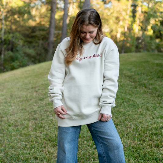 Woman wearing a cream sweatshirt with 'Barely Regulated' text in an outdoor setting